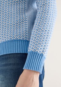 Manche de pull tricoté texturé bleu et blanc avec bord-côtes portée sur un jean en denim bleu, montrant une partie du bras et de la main d'une personne.