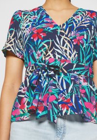 Blouse à manches courtes et col en V avec imprimé floral tropical en bleu, rose, rouge et vert, dotée d'une ceinture nouée à l'avant et d'un bas style peplum.