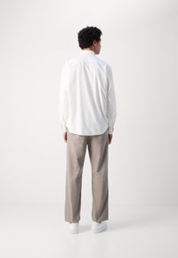 Camicia bianca a maniche lunghe con yoke posteriore, abbinata a pantaloni larghi grigi chiari e sneakers bianche, su uno sfondo neutro.