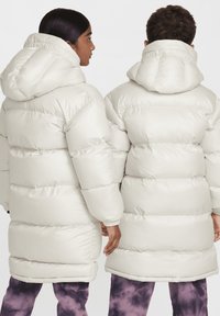 Dos personas visten chaquetas largas y acolchadas de color crema con capuchas, que presentan secciones acolchadas horizontales y puños elásticos.