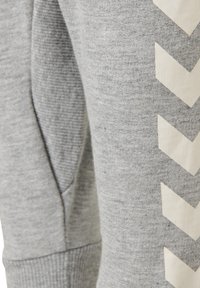 Grå sweatshirttyg med ribbad textur, med en sidoficka och ett stort vitt chevronmönster längs en ärm.