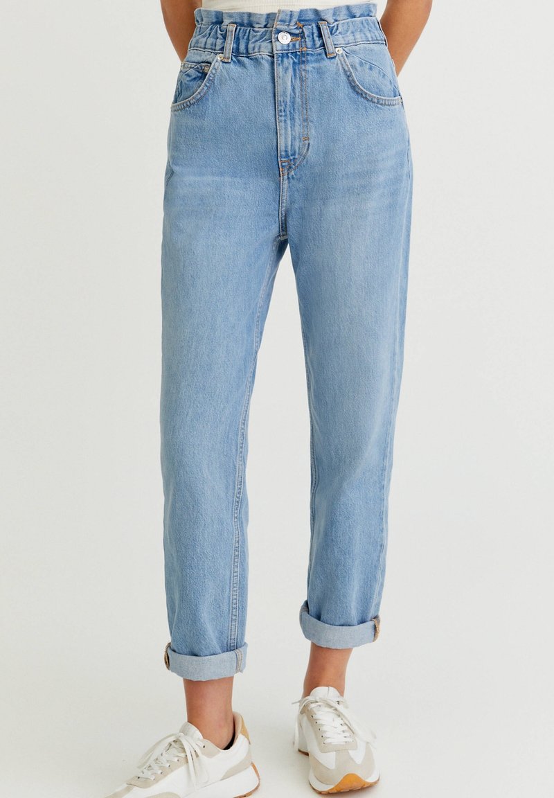 Jean taille haute bleu clair avec taille effet papier froissé et revers roulés, porté avec des baskets blanches sur un fond uni.