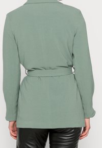 Blazer verde menta con cintura e maniche lunghe, tessuto liscio, collo a punta e taglio sartoriale, con un dettaglio di vita arricciata sul retro.