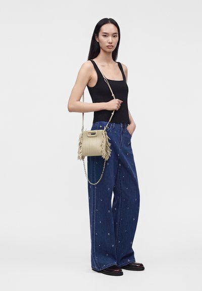 Jeune femme portant un haut noir sans manches et un jean large bleu à motifs, tenant un sac à bandoulière beige à franges avec une chaîne dorée.