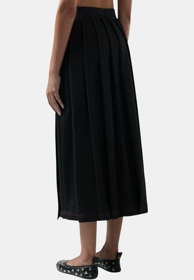 Vrouw die een zwarte geplooide midirok en zwarte ballerina's met studs draagt, staand met haar armen ontspannen langs haar zij.