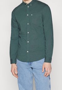 Camicia button-up verde scuro con colletto, maniche lunghe e tasca sul petto. Presenta bottoni bianchi e una texture di stoffa. Indossata con jeans azzurri chiari.