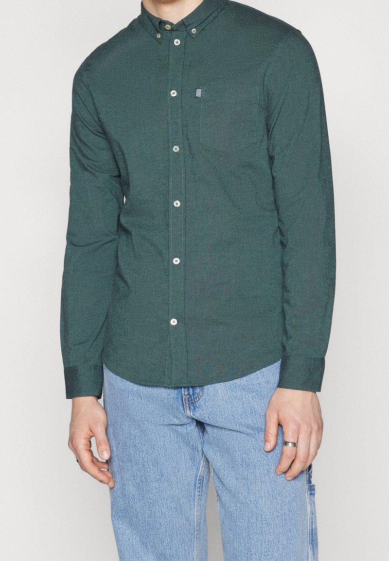 Camicia button-up verde scuro con colletto, maniche lunghe e tasca sul petto. Presenta bottoni bianchi e una texture di stoffa. Indossata con jeans azzurri chiari.