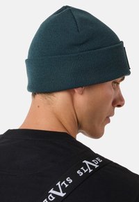 Cappello di lana teal con una texture a costine, presenta un risvolto e una corona piatta, indossato sulla testa di una persona con una maglietta nera visibile.
