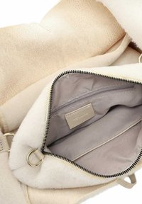 Beige-käsilaukku, jossa on pehmeä, teksturoidu ulkopinta. Sisällä on sileä, vaalea harmaa vuori ja vetoketjullinen tasku. Kultaista koristelun osia.