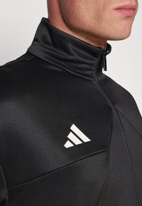 Blazer desportivo preto com gola alta, apresentando um fecho de zíper e o logótipo branco da Adidas. O material parece suave, com detalhes de design estruturados.