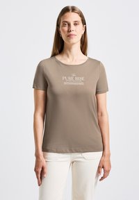 Žena s dlouhými světlými hnědými vlasy, oblečená v taupe tričku s bílým textem "The PURE RISE" a krémových kalhotách, stojící na pozadí čistě bílé stěny.