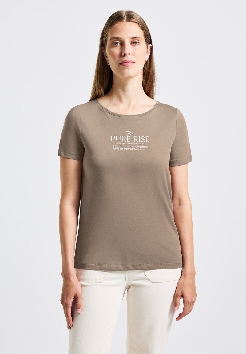 Žena s dlouhými světlými hnědými vlasy, oblečená v taupe tričku s bílým textem "The PURE RISE" a krémových kalhotách, stojící na pozadí čistě bílé stěny.