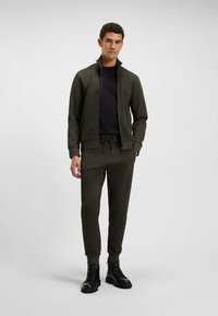 Donker olijfgroene trainingspak met ritsjack, lange mouwen en broek met koord. Gepaard met zwarte veterlaarzen. Matte textuur.