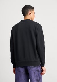 Svart sweatshirt med ribbad textur och långa ärmar, med en rundad halsringning och en avslappnad passform, kombinerat med lila tie-dye byxor.