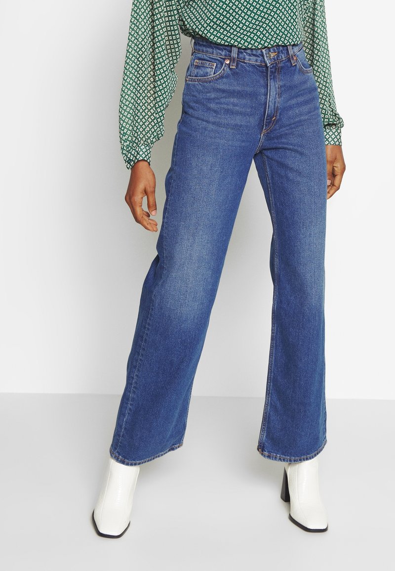 Jeans anchos de mezclilla azules, con cinco bolsillos, cintura alta y botas de tacón blancas. Textura suave con sutiles detalles de costura.