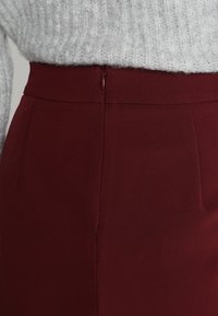 Jupe crayon bordeaux avec une texture lisse, dotée d'un zip discret à l'arrière et d'une taille épurée pour un look soigné.