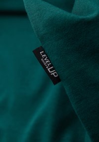 Tissu vert avec une étiquette noire de vêtement portant un texte blanc "LEVEL UP ombre" cousu près d'une couture.