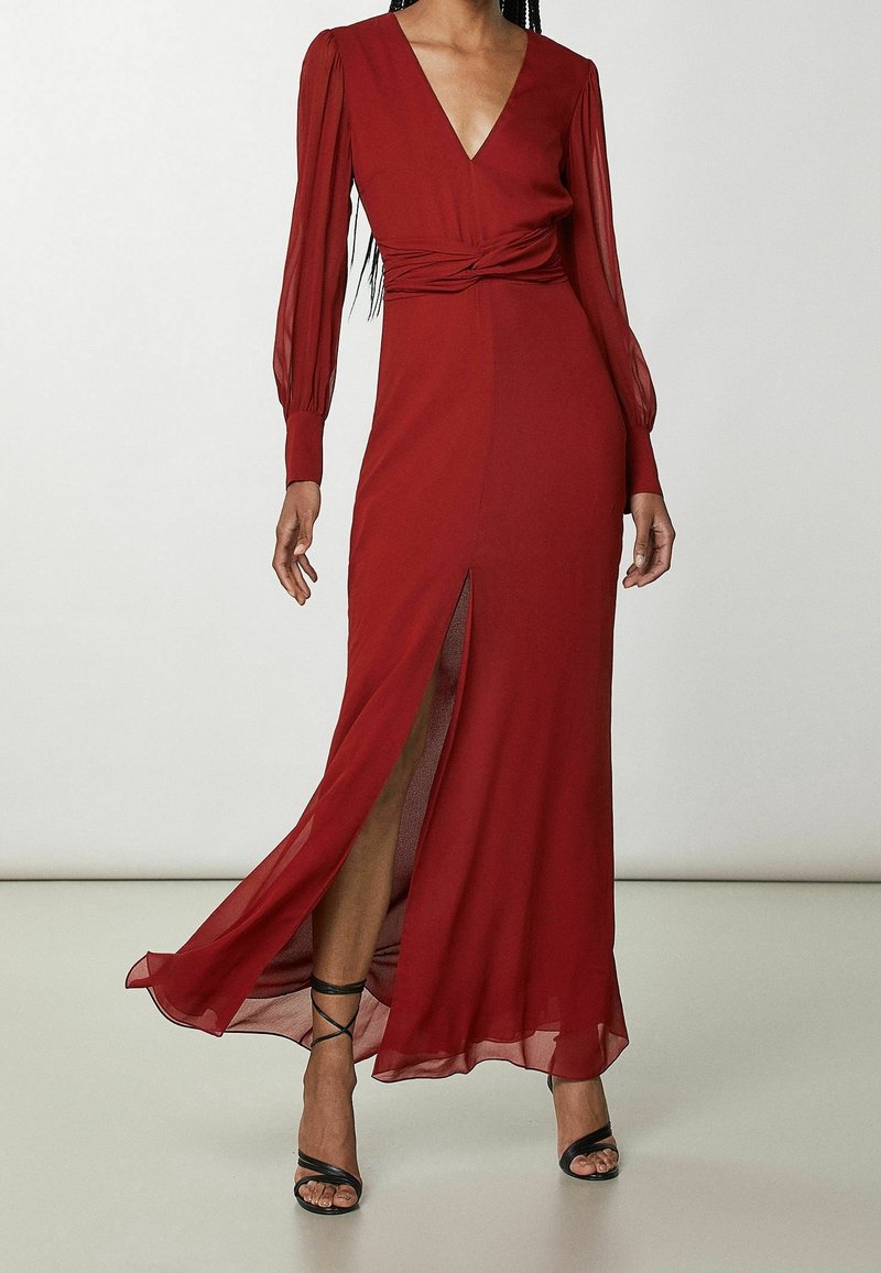 Femme portant une robe longue rouge foncé, avec des manches longues transparentes, un col en V, un détail torsadé à la taille, une fente à l'avant et des talons noirs à brides.