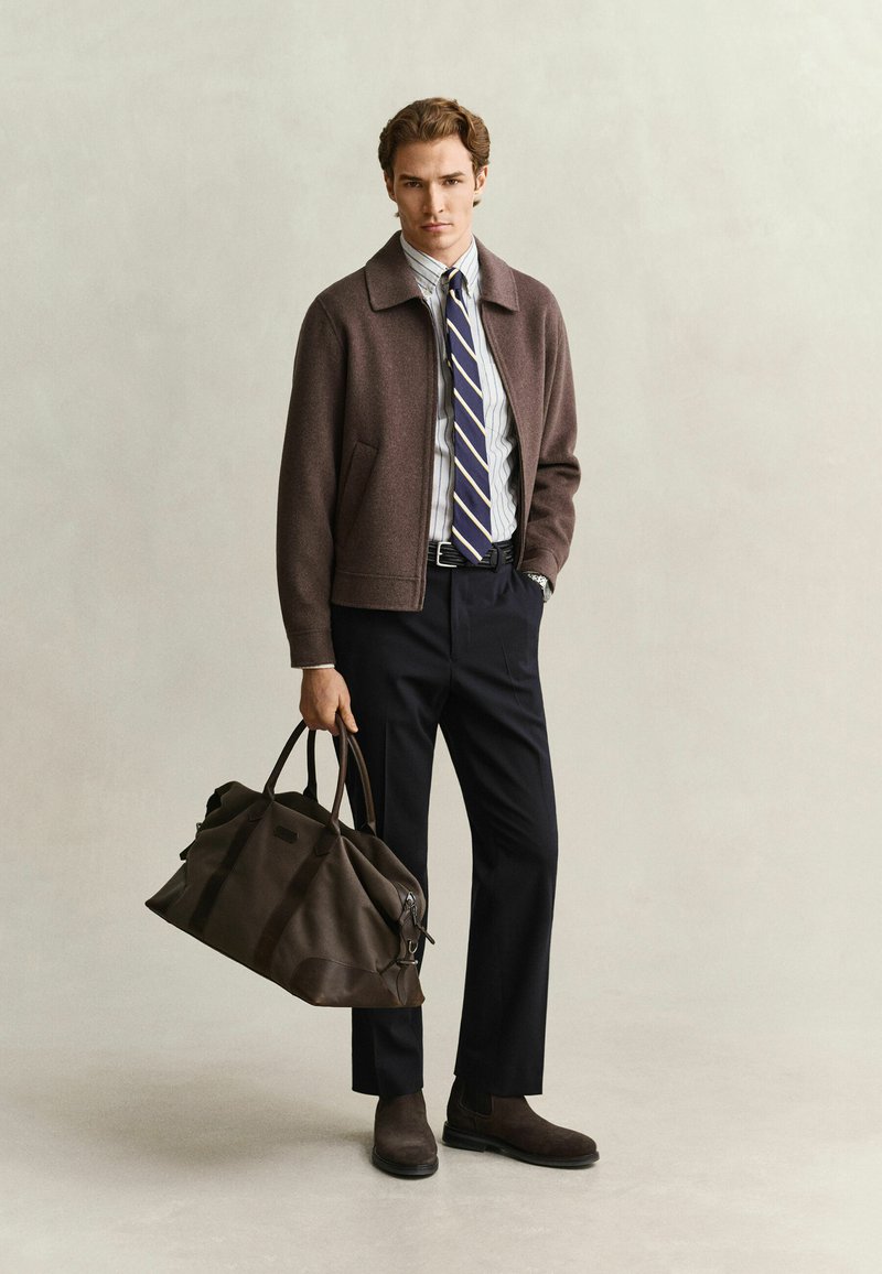 Homme en veste marron, chemise à rayures, cravate marine, pantalon noir et bottes marron tenant un grand sac de voyage en cuir marron, debout devant un fond uni.