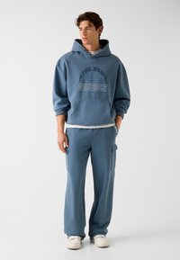 Niebieska bluza z kapturem i dopasowane spodnie typu cargo z nadrukami "ATHL.DET" i "GUESS". Tkanina wydaje się miękka, z luźnym krojem i kieszenią typu kangurka.