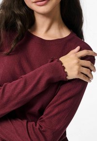 Bordo ilga rankovė su tekstūruotu audiniu, su raukinėtomis rankovėmis ir brūkšniuotu dizainu. Artimas vaizdas, kai rankos susikryžiuos ant krūtinės.