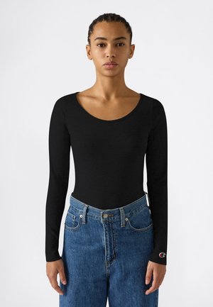 Vrouw met gevlochten haar die een aansluitend zwart langemouwen shirt en een high-waisted blauwe spijkerbroek draagt, staande tegen een effen witte achtergrond.
