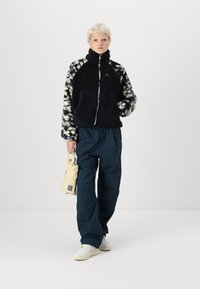 Chaqueta de forro polar negra con mangas de patrón multicolor, cremallera verde, combinada con pantalones azul oscuro y zapatillas blancas, sosteniendo una bolsa beige.