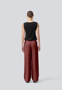 Schwarzes ärmelloses Top, rote weit geschnittene Hose mit glatter Textur, Rückansicht mit zwei Taschen, flache Schuhe und neutraler Hintergrund.