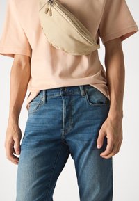 Beige tygmidjeväska som bärs över en ljusrosa t-shirt, kombinerad med mörkblå denimjeans. Mjuk textur och avslappnade designelement.