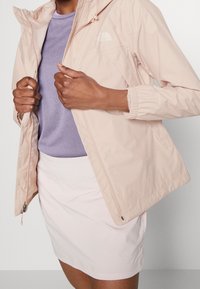 Giacca leggera rosa chiaro con cappuccio e polsini elasticizzati. Indossata sopra un top lavanda, abbinata a una gonne rosa pallido. Include dettagli con logo.