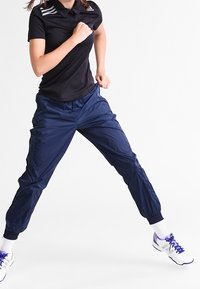 Schwarzes Polo-Shirt mit drei weißen Streifen auf der Schulter, kombiniert mit navyblauen Sporthosen. Das Modell nimmt eine dynamische Pose ein.
