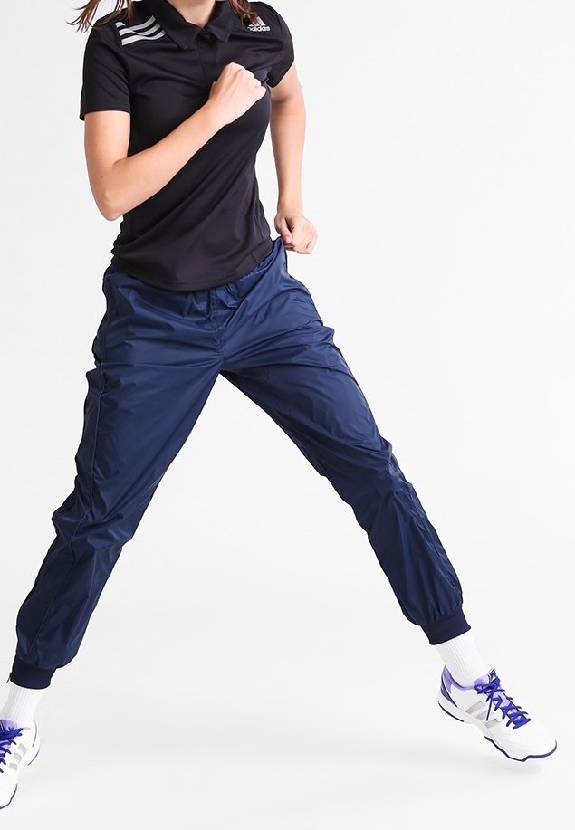 Schwarzes Polo-Shirt mit drei weißen Streifen auf der Schulter, kombiniert mit navyblauen Sporthosen. Das Modell nimmt eine dynamische Pose ein.