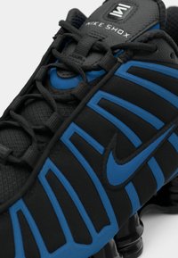 Close-up de um ténis Nike Shox preto e azul com cordões detalhados e elementos de sola almofadada visíveis.