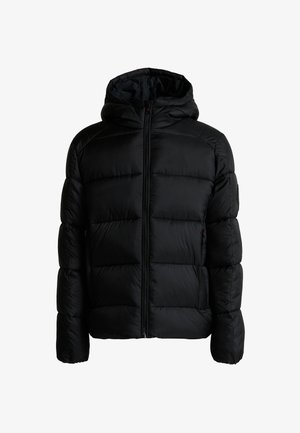 Staande jongen met gevlochten haar, een zwarte pufferjas, zwarte broek en zwart-rode sneakers tegen een effen grijze achtergrond.