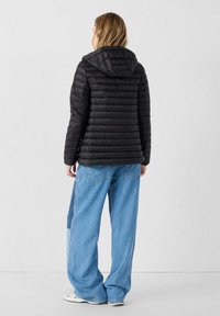 Schwarze Steppjacke mit Kapuze, mit horizontaler Steppung. Kombiniert mit hellblauen, weit geschnittenen Jeanshosen. Glatte Stoffstruktur.