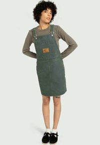 Denimova obleka v temno zeleni barvi z nastavljivimi naramnicami, sprednjim žepom in usnjenim nevihnikom. Nosimo jo čez sivo dolgosleeve majico in črne čevlje.