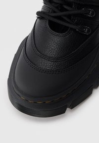 Prim-plan al vârfului unei bocanci de drumeție din piele neagră, cu un superior texturat, talpă groasă din cauciuc, șireturi negre și detalii cu cusături galbene.