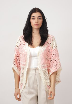 Vrouw met lang donker haar draagt een wit topje, beige broek en een roze-wit gehaakte omslagdoek, staand tegen een effen achtergrond.