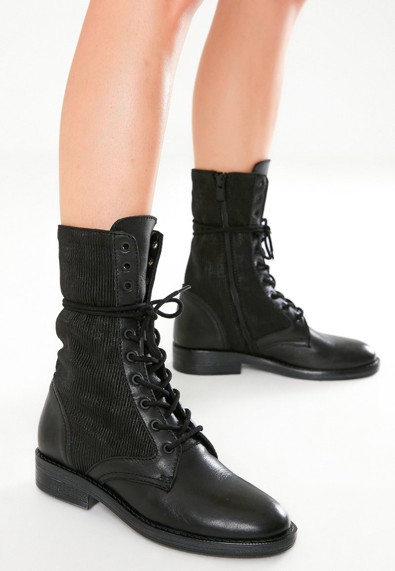 Bottes de combat en cuir noir à lacets sur le devant, panneaux latéraux texturés, fermetures éclair sur les côtés, portées sur des jambes nues sur un fond blanc uni.