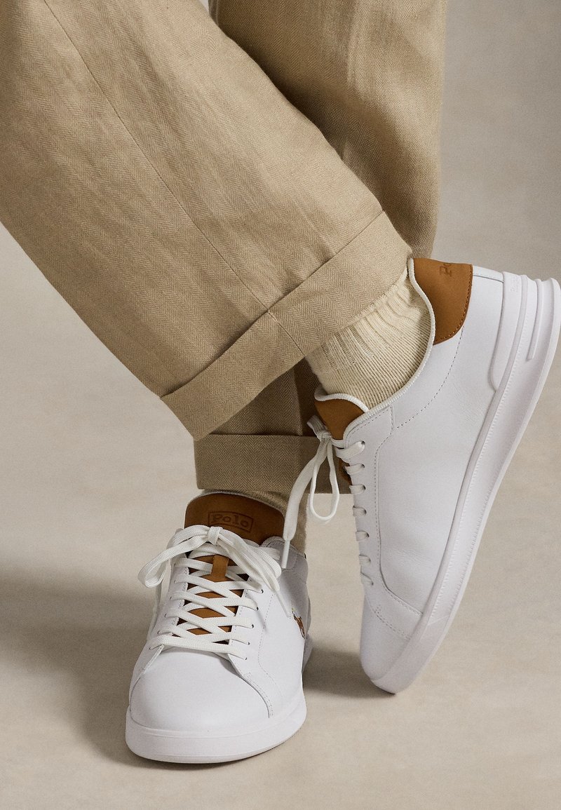 Personne portant un pantalon beige, des chaussettes crème et des baskets blanches avec des accents marron, un pied légèrement levé sur un fond neutre.