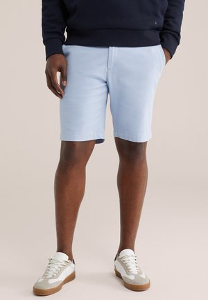 Mężczyzna w jasnoniebieskich szortach, granatowej bluzie i białych trampkach z beżowo-brązową podeszwą, stoi na jednolitym tle.