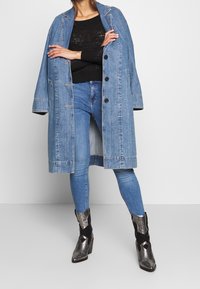 Płaszcz trencherowy z denimu, jasnoniebieski, z zapięciem na guziki, w oversize'owym kroju, z dwoma przednimi kieszeniami. Nosi się go na czarnej swetrze z dzianiny i dopasowanych niebieskich dżinsach, z metalicznymi botkami na kostkach.