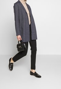 Harmaa pitkä bleiseri beigen poolopaidan, mustien kapenevien housujen ja mustien loafer-kenkien päällä, pitäen kädessään pientä mustaa käsilaukkua, jossa on teksturoitu pinta.