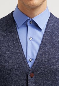 Mörkblå stickad cardigan med v-ringning, lagd över en ljusblå skjorta med knappar. Har mörka knappar och en strukturerad tyg.