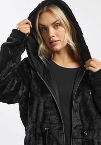 Hoodie in finta pelliccia nera con zip, dettagli con coulisse e tessuto morbido. Indossato sopra una maglietta a maniche lunghe nera.