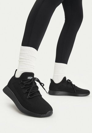 Osoba nosząca czarne dziergane trampki z czarnymi podeszwami, białe prążkowane skarpetki oraz czarne legginsy, stojąca na jasnym tle.