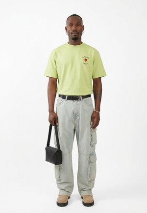 Uomo che indossa una t-shirt grafica verde chiaro, pantaloni cargo grigi, stivali beige, che tiene una borsa nera su sfondo bianco.