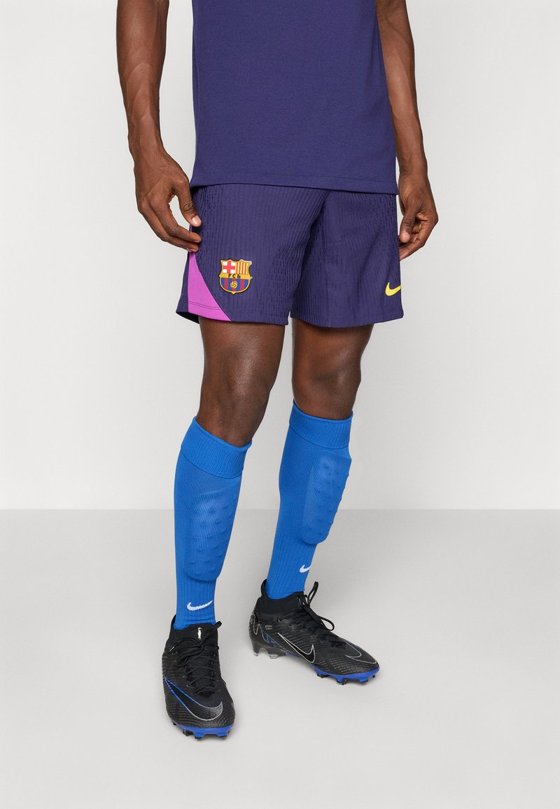 Moški nosi vijolične nogometne hlače FC Barcelona, modre zaščitne obloge za goleni Nike, črne nogometne čevlje Nike in stoji ob preprosti ozadju.
