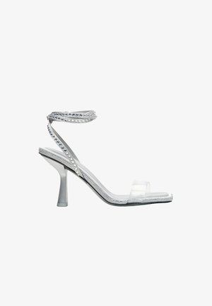 Průhledné sandály na podpatku s průsvitným svrškem, zdobené stříbrnými cvočky a jemným nártkem, umístěné na štíhlém podpatku.