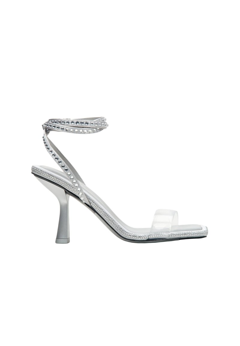 Transparante sandalen met een doorzichtig bovenwerk, versierd met zilveren studs en een delicaat enkelbandje, geplaatst op een slanke hak.